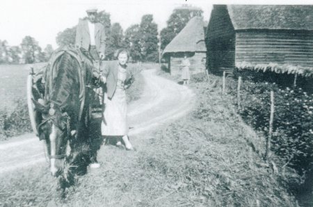 B&W photograph of a Lower Radley scence in 1937 featuring a horse and cart on a track, with a man standing on the cart and a young lady standing next to the cart; a barn and thatched cottage can be seen in the background (a lady is standing on front of the barn looking at the group on the track ahead of her)